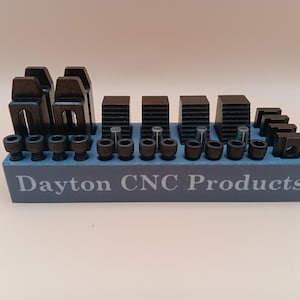 May include: A collection of black metal CNC products, including clamps, blocks, and bolts, arranged on a blue rectangular base. The base has the text "Dayton CNC Products" printed on it. The items are displayed against a neutral background.