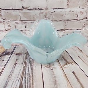 May include: A pale blue glass bowl with a ruffled, flower-like rim. The bowl features a smooth, translucent finish with a soft gradient of light blue color throughout the glass structure, creating a wide, open shape with curved, undulating edges.