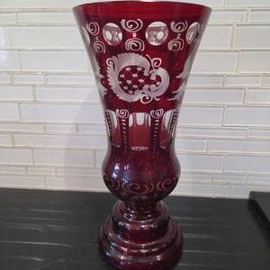 May include: A deep red glass vase featuring intricate white etched patterns. The design includes circular motifs near the flared rim, floral-inspired engravings in the center, and vertical panels above a rounded base, all crafted in cut-to-clear glass.