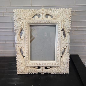 May include: A rectangular picture frame in an off-white, cream color. The frame features an intricate, carved relief pattern with scrolling leaf and floral motifs surrounding a central rectangular opening with a gray backing insert.