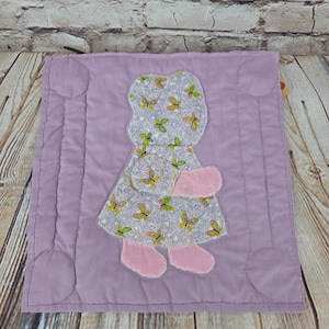 May include: A square lavender quilted fabric piece featuring a central applique of a figure wearing a dress with a white background and a pattern of small yellow and green butterflies. The figure has solid pink fabric hands and feet.