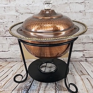 May include: A copper-colored chafing dish with a hammered texture, a clear crystal handle, and a silver-colored rim. The dish sits on a black metal stand with decorative scrollwork. The background is a white brick wall and wooden planks.