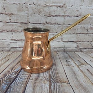 May include: A polished copper coffee pot with a wide base, a narrow neck, and a flared rim. The pot features a long, slender, gold-toned metal handle attached to the side with a single rivet. The interior of the pot is lined with silver-toned metal.