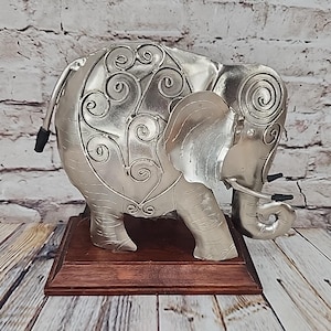 May include: A silver-colored elephant figurine with intricate scrollwork on its body, standing on a dark brown wooden base. The elephant has detailed features, including tusks and a raised trunk. The background features a brick wall and wooden planks.