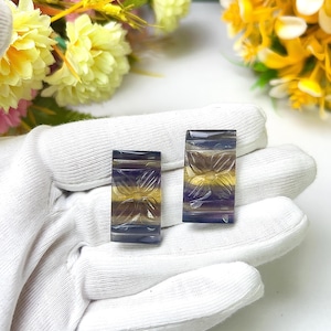 May include: Two rectangular, carved fluorite gemstones. The stones feature a floral design and are layered with bands of purple, yellow, and brown. The gemstones are held in a white gloved hand, with yellow and pink flowers in the background.