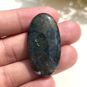 Könnte beinhalten: Ein oval geformter Edelstein mit polierter Oberfläche. Der Stein weist eine Mischung aus dunkelgrünen, blauen und weißen Farbtönen auf, wodurch ein marmorierter Effekt entsteht. Der Edelstein wird in einer Hand gehalten, was seine natürliche Schönheit und einzigartigen Farbvariationen hervorhebt.