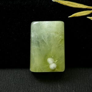 May include: A rectangular, polished gemstone cabochon in a pale, translucent lime green colour. The stone features internal radiating, feathery white inclusions and two distinct, small, opaque white spherical clusters near the bottom edge.