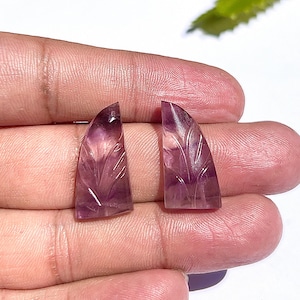 Könnte beinhalten: Zwei Amethyst-Edelstein-Cabochons, jeweils mit einem Blattmuster geschnitzt. Die Edelsteine haben eine durchscheinende violette Farbe mit sichtbaren inneren Mustern. Die Cabochons werden in einer Hand gehalten.
