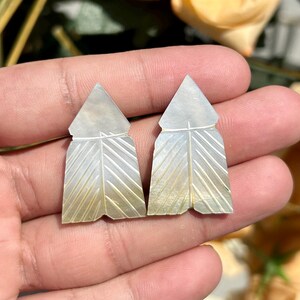 Könnte beinhalten: Zwei perlmuttfarbene, federförmige Ohrringe. Jeder Ohrring hat eine dreieckige Spitze und ein detailliertes, abgewinkeltes Federdesign. Die Ohrringe werden in einer Hand gehalten, mit einem unscharfen Hintergrund.