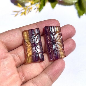 Könnte beinhalten: Zwei geschnitzte Edelsteinanhänger. Jeder Anhänger ist rechteckig mit einem floralen Design, das in die Oberfläche geschnitzt ist. Die Edelsteine sind eine Mischung aus Gelb-, Lila- und Brauntönen. Die Anhänger werden in einer Hand gehalten.