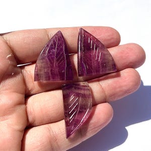 Könnte beinhalten: Drei polierte, violette Fluorite-Edelsteinblätter mit geschnitzten Blattaderdetails. Die Edelsteine werden in einer Hand vor weißem Hintergrund gehalten. Die Edelsteine sind von transluzenter violetter Farbe.