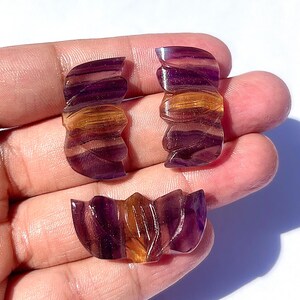 Könnte beinhalten: Drei geschnitzte Fluorite-Edelstein-Stücke. Die Stücke sind eine Mischung aus lila und goldfarbenen Farben. Die beiden oberen Stücke haben eine gebogene Form, und das untere Stück hat eine fächerartige Form. Die Edelsteine werden in einer Hand gehalten.