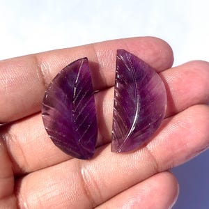 Könnte beinhalten: Zwei geschnitzte Blattanhänger aus Amethyst-Edelstein. Die Anhänger haben eine tiefviolette Farbe mit komplizierten Aderdetails. Die Edelsteine werden in einer Hand gehalten, wodurch ihre polierten Oberflächen zur Geltung kommen.