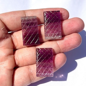Könnte beinhalten: Drei rechteckige, violette Fluorit-Edelsteine mit rautenförmiger Oberfläche. Die Steine werden in einer Hand gehalten, wodurch ihre polierte Oberfläche und der Farbverlauf von tiefem Violett zu durchscheinend hervorgehoben werden.