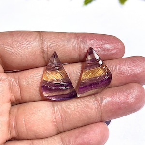 May include: Two polished, triangular gemstones with a layered design. The stones feature bands of purple, gold, and clear hues. The gemstones are held in a hand, showcasing their intricate details and natural beauty. The overall aesthetic is elegant and refined.