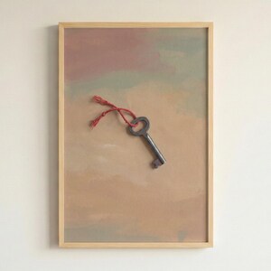 Puede incluir: Obra de arte enmarcada que presenta una llave antigua con un cordón rojo sobre un fondo abstracto apagado de tonos beige, rosa y verde. La llave es metálica y la obra está en un marco de madera clara.