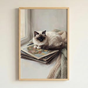 May include: A framed wall art print featuring a painting of a fluffy Birman cat with dark brown points resting on a stack of books near a window. A soft, textured, light-colored knit blanket is draped beside the books in the composition.