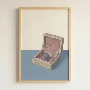 May include: A framed painting of a small, open wooden box with a pink interior, containing a silver thimble. The box sits on a blue surface, with a cream-colored background above. The artwork is displayed in a natural wood frame.