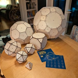 Puede incluir: Una colección de seis esferas poliédricas geométricas de madera clara de varios tamaños, cada una con puntos de conexión oscuros en los vértices. Se incluyen dos tarjetas de instrucciones azules con el texto TSIGILI BUM y diagramas geométricos.