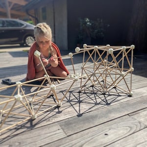 May include: A construction toy set featuring thin wooden rods connected by small, off-white geometric joints. The pieces are assembled into a complex, three-dimensional lattice structure with triangular and square patterns, resting on a wooden surface.