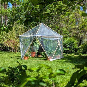 Puede incluir: Un invernadero de jardín hexagonal con estructura de madera y paneles de plástico transparente. La estructura está situada sobre un césped y contiene varias plantas en macetas y una silla colgante negra en su interior.