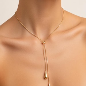 May include: A gold-colored necklace with a delicate chain and a sliding pendant design. The pendant features two teardrop-shaped charms that hang at different lengths. The necklace is worn around the neck.