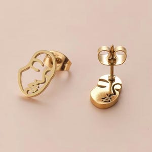 May include: A pair of gold-colored stud earrings. Each earring features a minimalist face design. One earring is a cutout, while the other is a solid shape with a face etched into the metal. The earrings are displayed on a light pink surface.