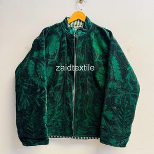 Könnte beinhalten: Eine dunkelgrüne Samtjacke mit aufwendiger, tonaler Stickerei aus Blumen- und Blattmustern. Die Jacke hat einen Stehkragen, einen zentralen Reißverschluss und lange Ärmel. Der Text "zaidtextile" ist in der Mitte aufgedruckt.