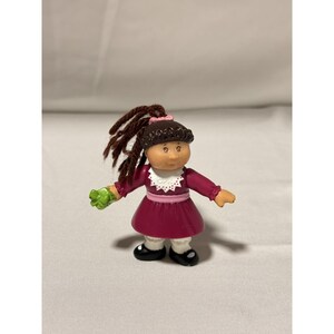 Può includere: Una piccola bambola in plastica con capelli in lana marrone scuro raccolti in una coda di cavallo, che indossa un vestito bordeaux con colletto in pizzo bianco, una fascia rosa in vita, leggings bianchi con volant e scarpe nere.