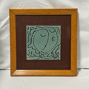 May include: Framed artwork depicting a teal face with black line art on a brown background. The artwork is set within a square, light brown wooden frame. The image has a minimalist, abstract style.