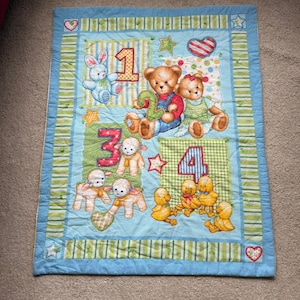 May include: A rectangular baby quilt featuring a light blue border with green striped accents. The quilt displays four numbered sections with illustrations of a blue rabbit, two teddy bears, three white lambs, and four yellow chicks with hearts and stars.
