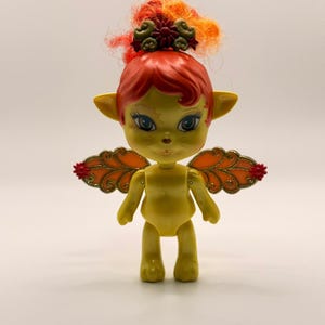 May include: A yellow doll with pointed ears, large dark eyes, and a small nose. It has bright red hair styled with a dark red and gold floral hair accessory. The doll features orange, leaf-shaped wings with gold glitter outlines and red accents.