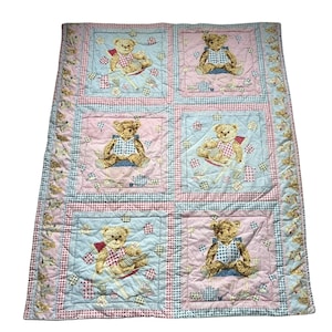 May include: A quilted blanket featuring a grid of six square panels, each displaying a teddy bear sitting in a chair. The panels alternate between light blue and pink backgrounds, surrounded by a border of small teddy bear illustrations.