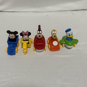 Pode incluir: Um conjunto de cinco veículos de brinquedo de plástico com personagens da Disney. Mickey Mouse num comboio azul, Minnie Mouse numa mota amarela, Pateta num barco vermelho, Pluto num carro laranja e Pato Donald num avião verde.