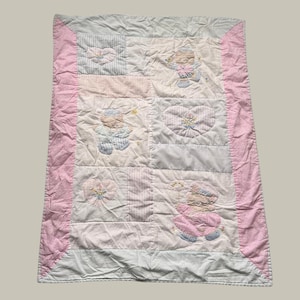 May include: A rectangular baby quilt featuring a patchwork design with a pink border. The quilt includes white, light blue, and pink fabric panels with embroidered motifs of teddy bears, hearts, and floral patterns in soft pastel colour tones.