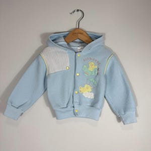 May include: A light blue hooded jacket with long sleeves and ribbed cuffs. The front features yellow snap buttons, a white textured panel on the right chest, and embroidered yellow bears with a green aeroplane and parachute, plus the text AIRBORNE.