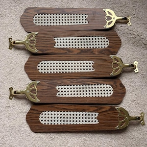 May include: Five ceiling fan blades featuring a dark brown wood grain finish with a central rectangular cutout filled with a white woven cane mesh pattern. Each blade is attached to a polished gold-tone metal bracket with a decorative design.