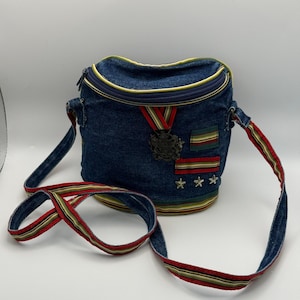 May include: A dark blue denim crossbody bag with a rounded top zipper closure. The bag features a decorative metal crest on a red, yellow, and green ribbon, two horizontal striped fabric patches, and three small silver-toned star-shaped metal studs.