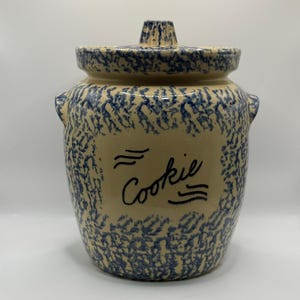 May include: A ceramic cookie jar with a cream-coloured base and a blue sponge-painted pattern. The jar features two side handles, a matching lid with a central knob, and the word Cookie written in black cursive script on the front surface.