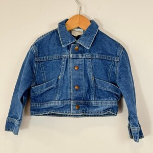 May include: A medium-wash blue denim jacket featuring a classic collar, long sleeves with buttoned cuffs, and a front button closure with four metallic buttons. The jacket has two horizontal seam details across the chest and a cropped hemline.