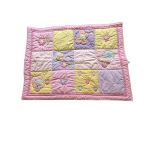 May include: A rectangular quilted blanket featuring a patchwork design of pink, yellow, and lavender squares. The quilt is decorated with appliqued butterflies and small flowers in various pastel colours, finished with a solid pink border.