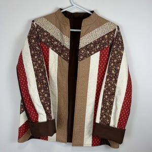 Peut inclure: Une veste matelassée présentant un motif à chevrons et à rayures verticales dans des tons de marron, crème et rouge. Le vêtement possède un col officier, des manches longues avec des poignets marron unis et une doublure intérieure marron.