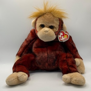 May include: A plush orangutan toy with dark reddish-brown fur on the body and limbs, and lighter tan fur on the face, hands, and feet. The toy features a tuft of spiky orange hair on its head, black eyes, and a red heart-shaped Ty tag attached.