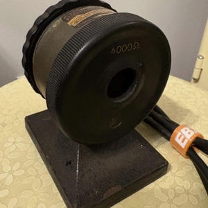 May include: A vintage, industrial-style device with a dark olive-green cylindrical body and a black, ridged top. The device is mounted on a dark, square base. The number "4000Ω" is printed on the side. Black cables are attached.