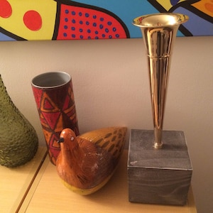 May include: A gold metal vase with a round top and a square base. The vase is sitting on a wooden table next to a brown and orange ceramic bird figurine, a green glass bottle, and a ceramic vase with a geometric pattern.