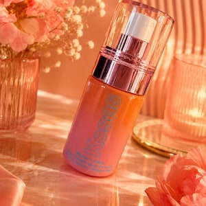 Op de afbeelding: Een cosmeticafles met een kleurovergang van oranje naar roze, met de merknaam "Kosas" in blauw. De fles heeft een transparante pomp en dop, met een metallic ros&eacute;gouden accent. Het product is gelabeld "Plump + Juicy".