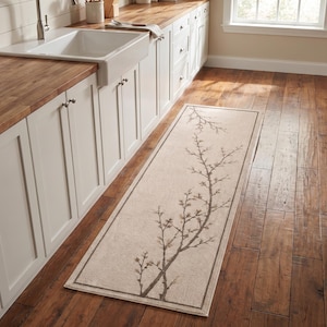 Puede incluir: Una alfombra de pasillo rectangular con fondo beige claro y un patrón de ramas botánicas en color marrón oscuro. La alfombra tiene un borde fino de color marrón oscuro y está colocada sobre un suelo de madera en una cocina con armarios blancos.