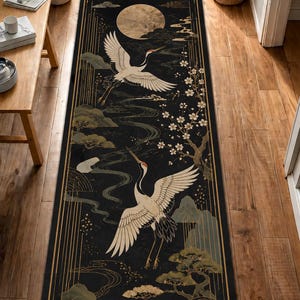 Puede incluir: Una alfombra rectangular tipo pasillero con fondo negro y estampado gráfico en dorado y blanco. El diseño muestra dos grullas blancas en vuelo, una luna llena grande, pinos estilizados y ramas florecidas enmarcadas por bordes geométricos dorados.