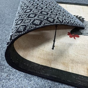 Pu&ograve; includere: Un tappeto rettangolare con superficie beige caratterizzato da un'illustrazione di un gallo bianco e nero con cresta rossa. Il tappeto &egrave; ripiegato per rivelare un retro nero con un motivo geometrico a rombi bianchi e una trama antiscivolo.