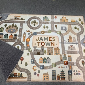 May include: A rectangular play rug featuring a printed town map with roads, buildings, trees, and a train. The center displays the text JAMES TOWN. The rug has a light-colored top surface with a detailed city graphic and a dark, patterned underside.
